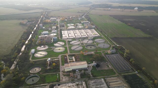 Aerial view of a wastewater treatment plant with multiple large circular clarifier tanks, sedimentation basins, channels and filtration structures arranged in a geometric industrial water-processing c