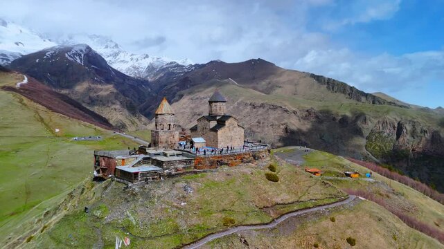 Aerial view of Gergeti Trinity Church with people on the terrace against snow-capped mountains in Mtskheta-Mtianeti, Georgia.