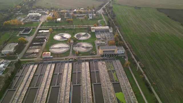 Aerial view of a large wastewater treatment plant with circular settling tanks, rectangular basins, pumping facilities, and green service roads set among agricultural fields and a canal under soft ove