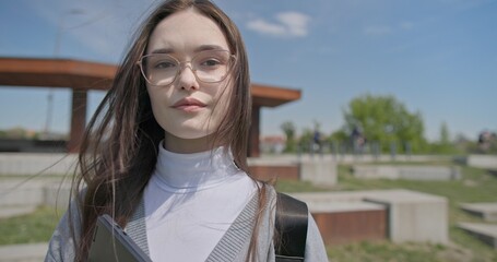 A hand-held portrait of a young female student in casual clothes, wearing glasses and a backpack, holding a notebook and laptop in her hands and posing looking at the camera while standing in a park  © Anatoly Repin