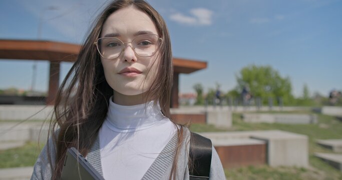 A hand-held portrait of a young female student in casual clothes, wearing glasses and a backpack, holding a notebook and laptop in her hands and posing looking at the camera while standing in a park 