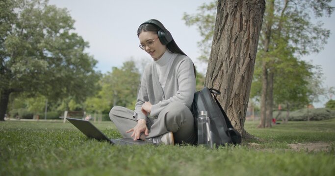 A hand-held shot of a young beautiful female student in casual clothes, wearing glasses and headphones, sitting on the grass under a tree in a city park and using a laptop while typing on the keyboard