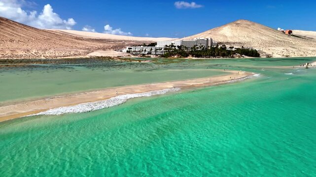Aerial view of Playa del Matorral featuring a sandbar and turquoise lagoon with a hotel complex and arid hills in the background, Canary Islands, Spain.