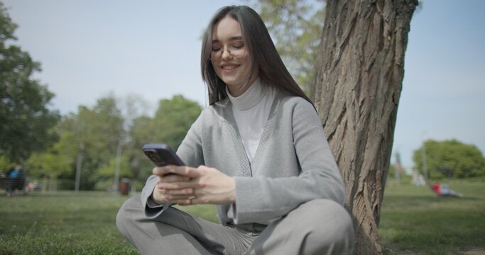 The shot is handheld and shows a young female student in casual clothes wearing glasses sitting on the grass under a tree in a park and using a mobile phone.