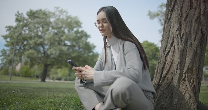 The shot is handheld and shows a young female student in casual clothes wearing glasses sitting on the grass under a tree in a park and using a mobile phone.