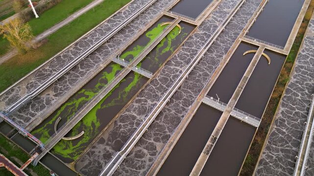 Aerial view of a large wastewater treatment plant with circular settling tanks, rectangular basins, pumping facilities, and green service roads set among agricultural fields and a canal under soft ove