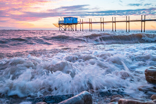 Cabane de p&ecirc;cheur sur pilotis au coucher du soleil, vagues et &eacute;cume sur les galets au premier plan, &agrave; Yves en Charente-Maritime - France