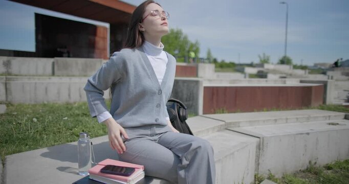 A handheld shot of a young female student in casual clothes sitting on a concrete bench in an urban park, enjoying a sunny summer day.