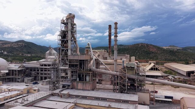 Aerial view of a large cement plant featuring a multi-stage cyclone preheater tower, clinker storage dome, vertical silos, rotary kiln and conveyor systems set against mountain backdrop
