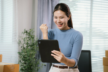 Happy young Asian female entrepreneur celebrating online business success while using tablet in home office surrounded by shipping boxes. © Kieng