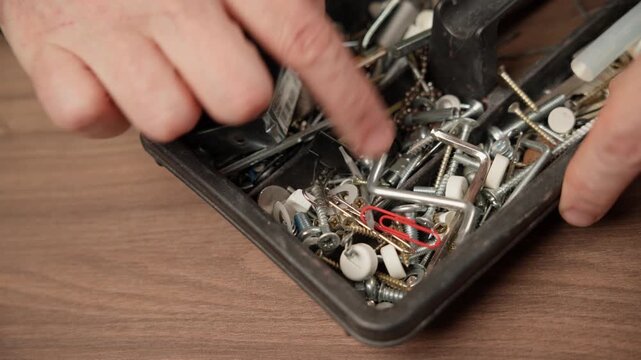 Handyman looking for a specific screw in a cluttered plastic compartment tray on a wooden table.
