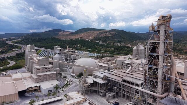 Aerial view of large cement plant featuring multi-stage cyclone preheater tower, spherical clinker storage dome, vertical silos, rotary kiln and conveyor systems surrounded by green hills