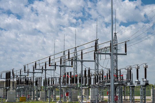 complex high-voltage equipment, including ceramic insulators and switchgear, at an electrical power substation under a cloudy blue sky.