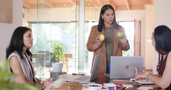 Diverse female coworkers watching presenter holding two apples showing choice at table with laptop
