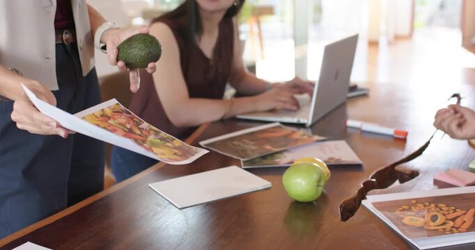 Diverse women seeing lead picking avocado and salad brochure, pitching recipe at table, copy space