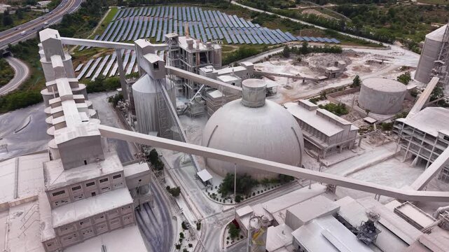 Overhead view of a large cement plant featuring a prominent spherical clinker dome, vertical silos, conveyor belts, grinding mills, and a solar panel array in the background