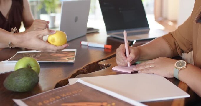 Diverse female coworkers discussing left woman picking lemon typing on laptop for notes at table