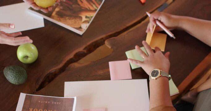 Two women planning recipe shoot on liveedge table, arranging cookbook fruit, notes, copy space