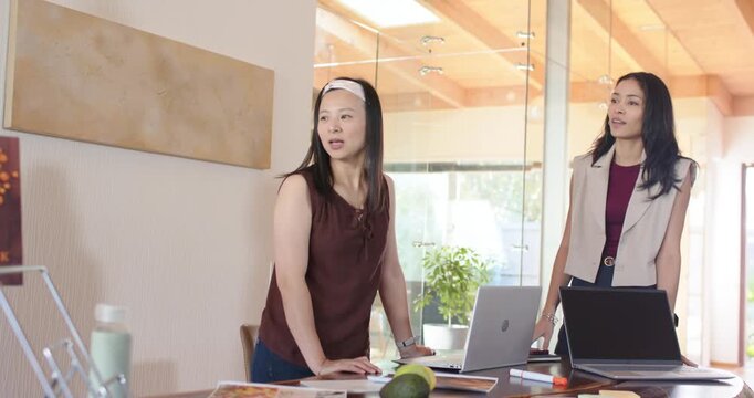 Diverse female colleagues standing at meeting table receiving brochure and reviewing laptop designs