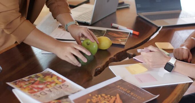 Diverse female coworkers styling avocado and apples left placing avocado right writing layout notes
