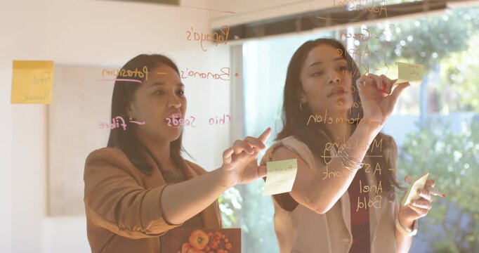 African American women planning at glass, left reaching, right writing with Postits in businesswear