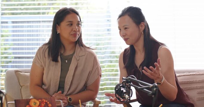 Diverse women podcasting, right narrating, fixing mic, left reaching bag smiling at home
