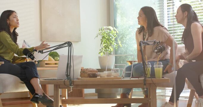 Diverse female hosts holding notes, gesturing at mics while recording in lounge around coffee table