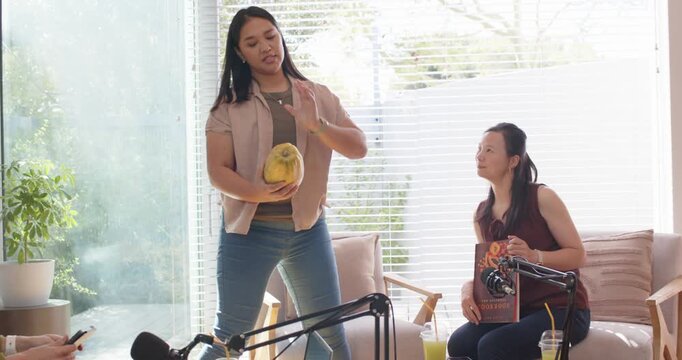 Diverse female friends podcasting at home, standing woman moving to center holding papaya, showing
