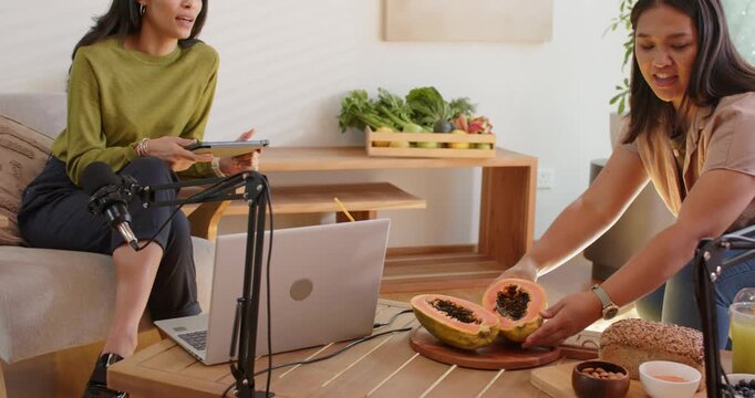 Diverse women sitting in studio, host near mics, guest approaching with papaya, placing for podcast