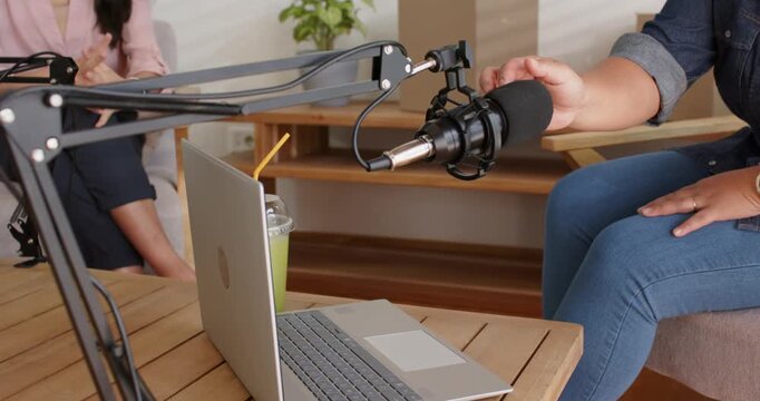 Diverse women hosts in studio adjusting mic after arm move, typing on laptop preparing to record