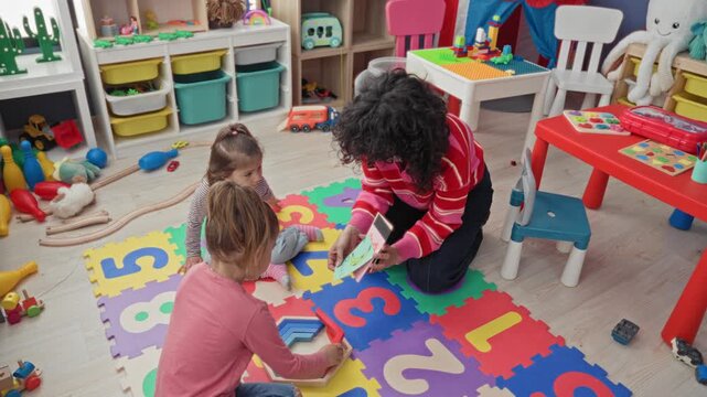 Woman kneeling showing flashcard to two girls playing with number foam mat and wooden blocks in a classroom building; playful learning interaction.