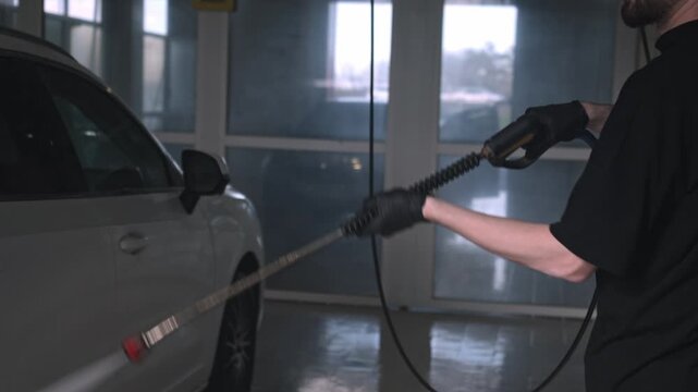 White Man Rinsing Car Front Fender With Pressure Lance In Dim Wash Bay, Deliberate Strokes Remove Road Salt