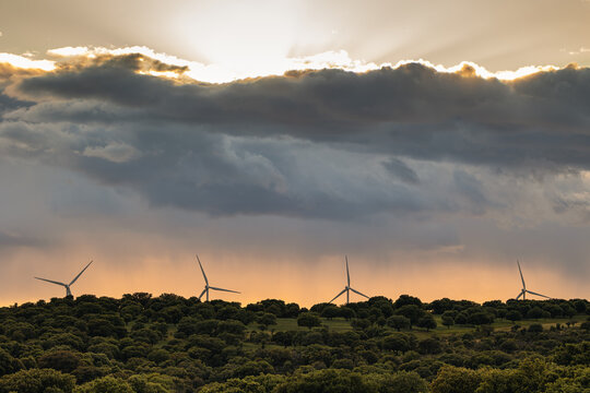 Wind energy turbines generating clean power on a hill, dramatic sunset clouds with sun rays breaking through sky