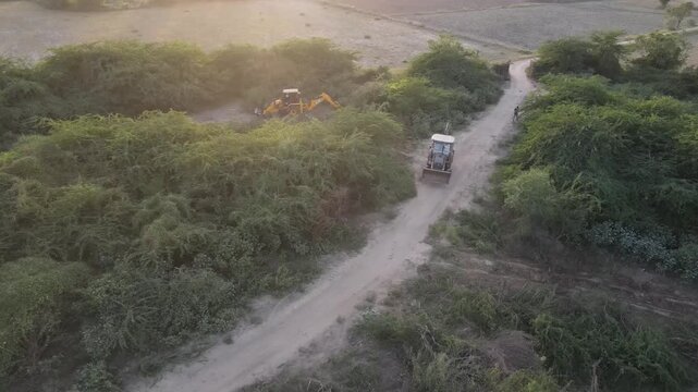 Cinematic drone footage of backhoe loaders clearing dense bushes along rural dirt roads during golden hour, showcasing land maintenance, vegetation removal, and countryside infrastructure development.