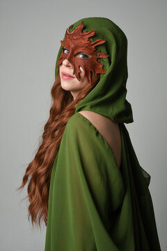 fantasy close up portrait of beautiful female model wearing leaf mask and green hooded cloak,  historical pagan fae  halloween costume. isolated figure on studio background.