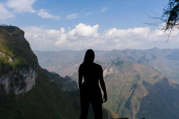 Silhouette stands atop cliff, gazing at vast mountain range. Clouds drift across bright blue sky above. Treetops frame the edge of the rugged landscape. Distant peaks fade into soft, atmospheric haze © jinhongljh