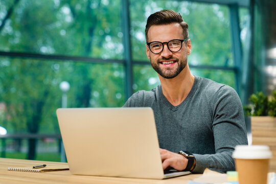 Professional man looking at camera using laptop in cozy coworking cafe, digital nomad working remotely in modern office space, modern career and flexible lifestyle concept