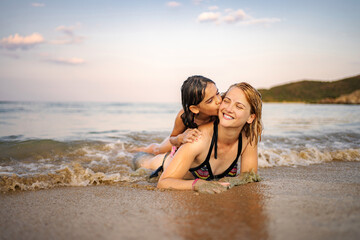 Fototapeta premium Mother and daughter smiling, lying in waves on a sandy beach