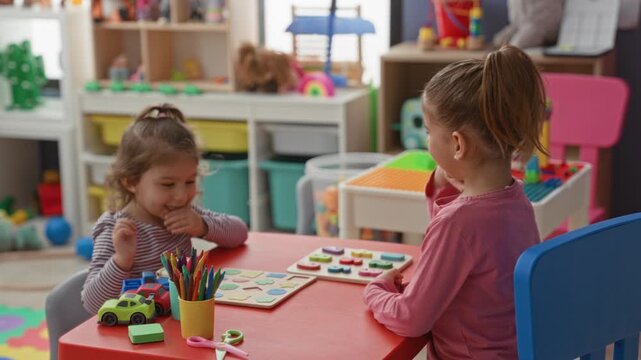 Two girls playing with wooden puzzles and toy cars at a small table, one raising hand for a high five in a daycare building; playful learning.
