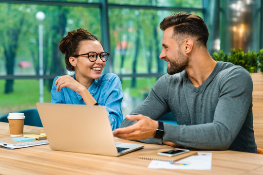 Two young Caucasian colleagues discussing and working together at office desk, using laptop to collaborate on a project in a modern and professional workspace