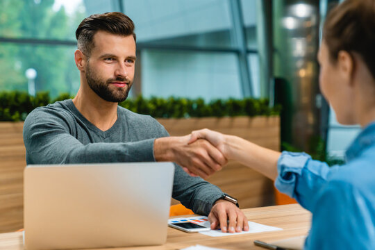 Two young business partners shaking hands at the office desk, sealing a deal or agreement in a professional and collaborative workspace.