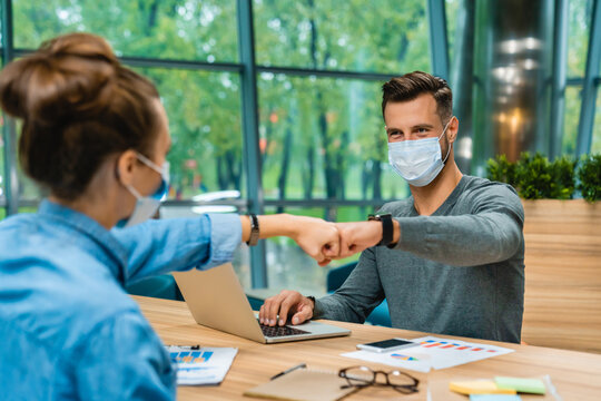Two young Caucasian colleagues wearing medical masks, joining fists in a gesture of teamwork and collaboration while working together in a modern office environment.