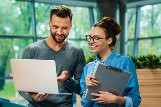 Two smiling Caucasian colleagues looking at a laptop together during a discussion in the office, collaborating and sharing ideas in a positive and engaging work environment.