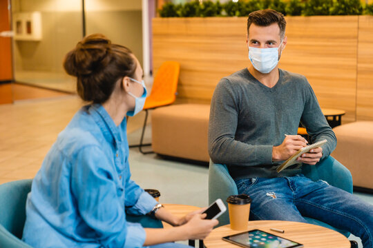Two young colleagues wearing medical masks, talking during their coffee break in the office, maintaining safety while enjoying a casual conversation in a professional environment.