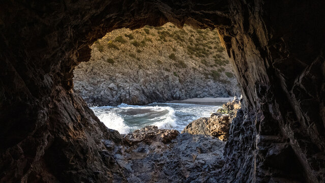 View of Spiaggia di Cala Domestica through a dark sea cave opening with turquoise waves crashing against rugged cliffs Provincia del Sulcis Iglesiente, Sardegna, Italy.