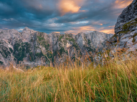 Rugged alpine landscape with wild grass in the foreground and towering mountain peaks under a dramatic sunset sky, inspiring outdoor adventure and tourism at Mangart, Italy-Slovenia border.