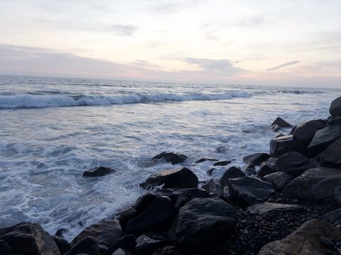 View of waves crashing against a rocky shoreline under a soft sunset sky Oceanside, California, United States.
