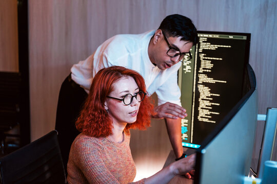 Two colleagues collaborate at a workstation, reviewing code on large monitors. One guides while the other types, focusing on software development and debugging in a modern office.