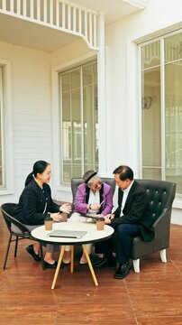 Three professionals meet on a patio, reviewing documents over coffee at a small round table beside a white building with large windows.