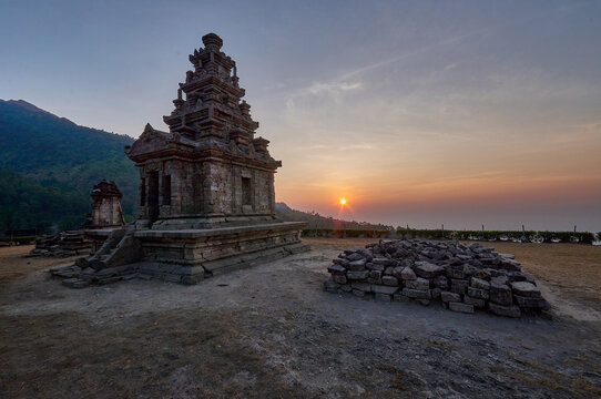 View of the ancient Gedong Songo Temple complex with stone ruins and a mountain backdrop during an orange sunset in Bandungan, Jawa Tengah, Indonesia.